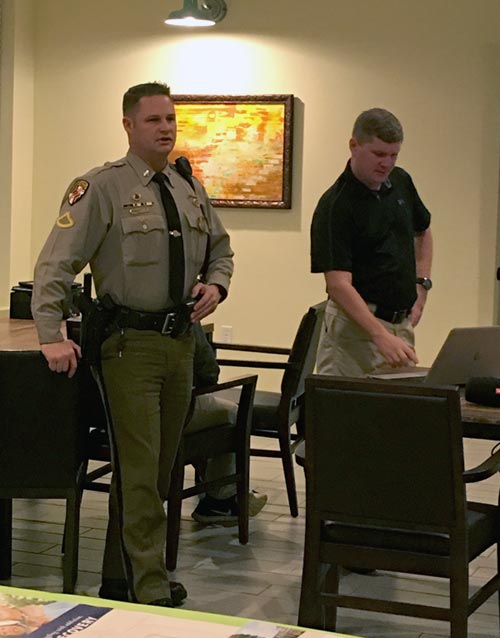 A police office standing in room speaking with another man next to him on a laptop